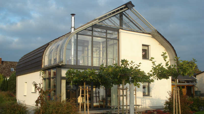 Wei&szlig;es Bio-Solar-Haus mit gebogener Glasfassade (Haus-im-Haus), Garten und Himmel bei Abendlicht.
